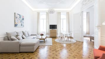 A spacious living room with a large L-shaped sofa, a coffee table, and a TV. The room has wooden parquet flooring and white walls.