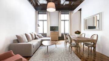 A modern living room with a sofa, a dining table, and chairs. There is a large window, a ceiling light, and a mirror on the wall.