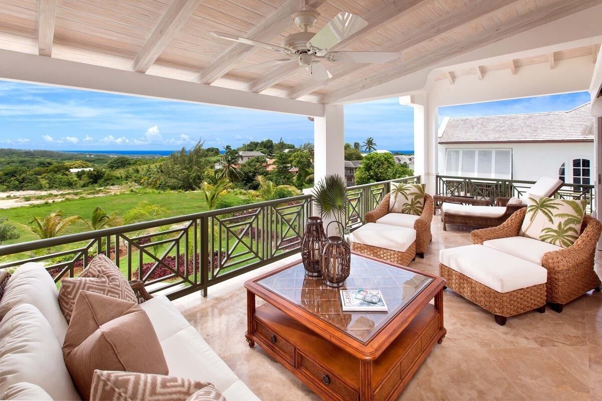 A spacious balcony with wicker furniture, a glass coffee table, and a view of a lush landscape.