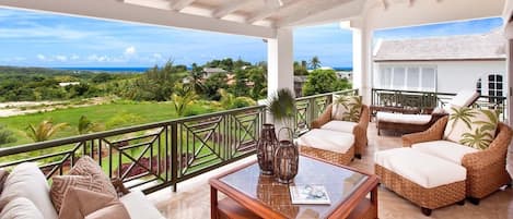 A spacious balcony with wicker furniture, a glass coffee table, and a view of a lush landscape.