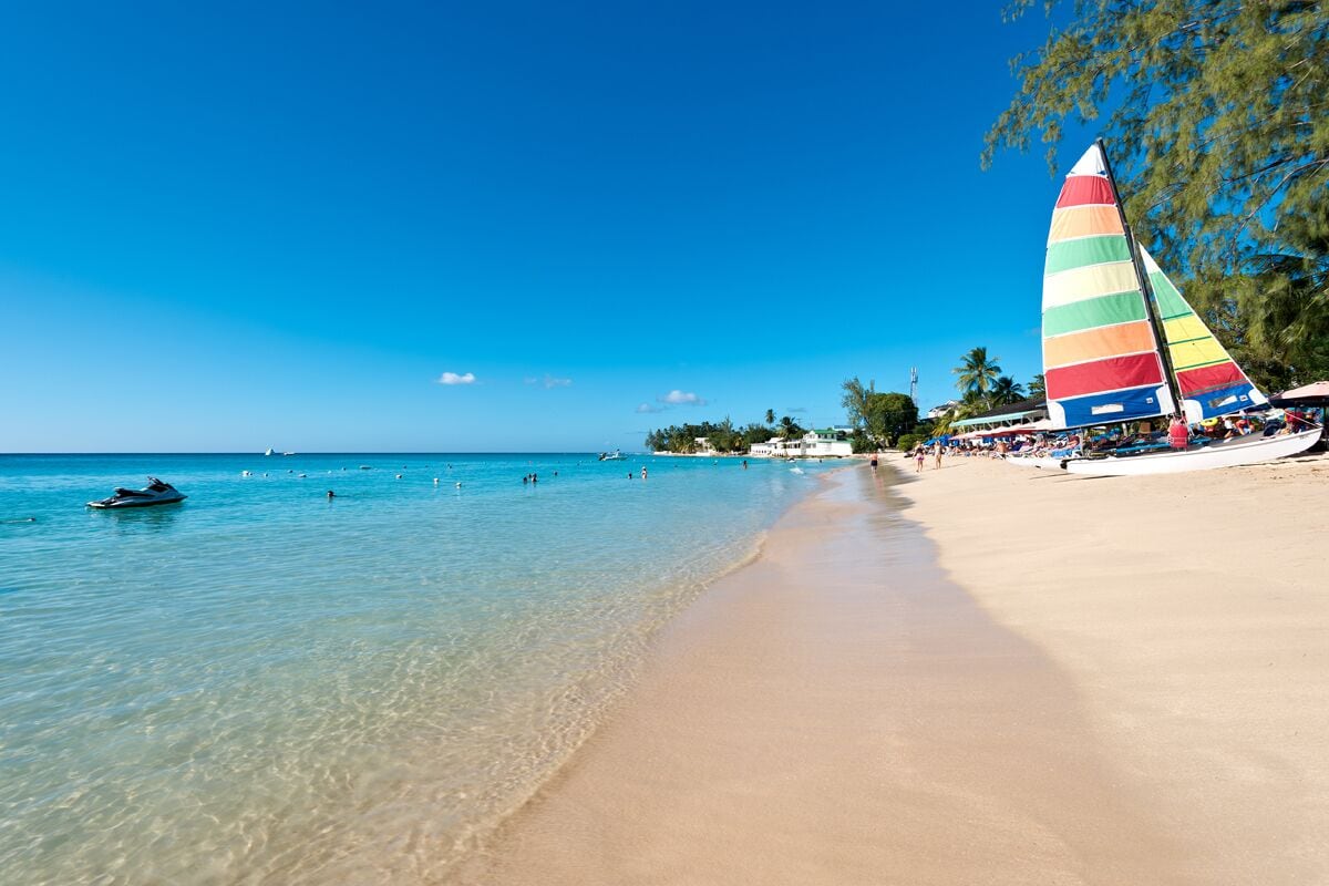 Ein Strand mit klarem türkisblauem Wasser, einem sandigen Ufer und einem farbenfrohen Segelboot.