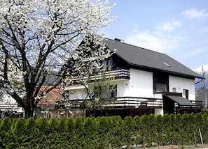 Une maison à deux étages avec un balcon, entourée d’une haie et d’un arbre en fleurs.