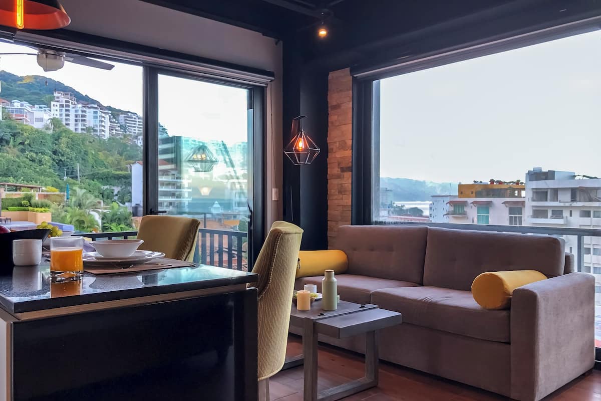 Un salon moderne avec un canapé, une table à manger et une vue sur le paysage urbain, grâce à de grandes fenêtres.