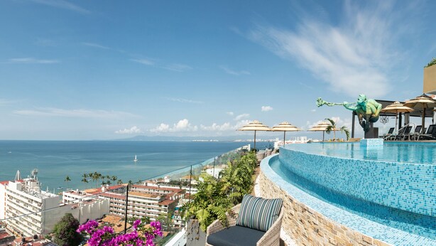 Une vue sur la piscine avec une vue dégagée sur la mer, un espace de détente avec des parasols, et un bâtiment doté d’un balcon donnant sur l’océan.