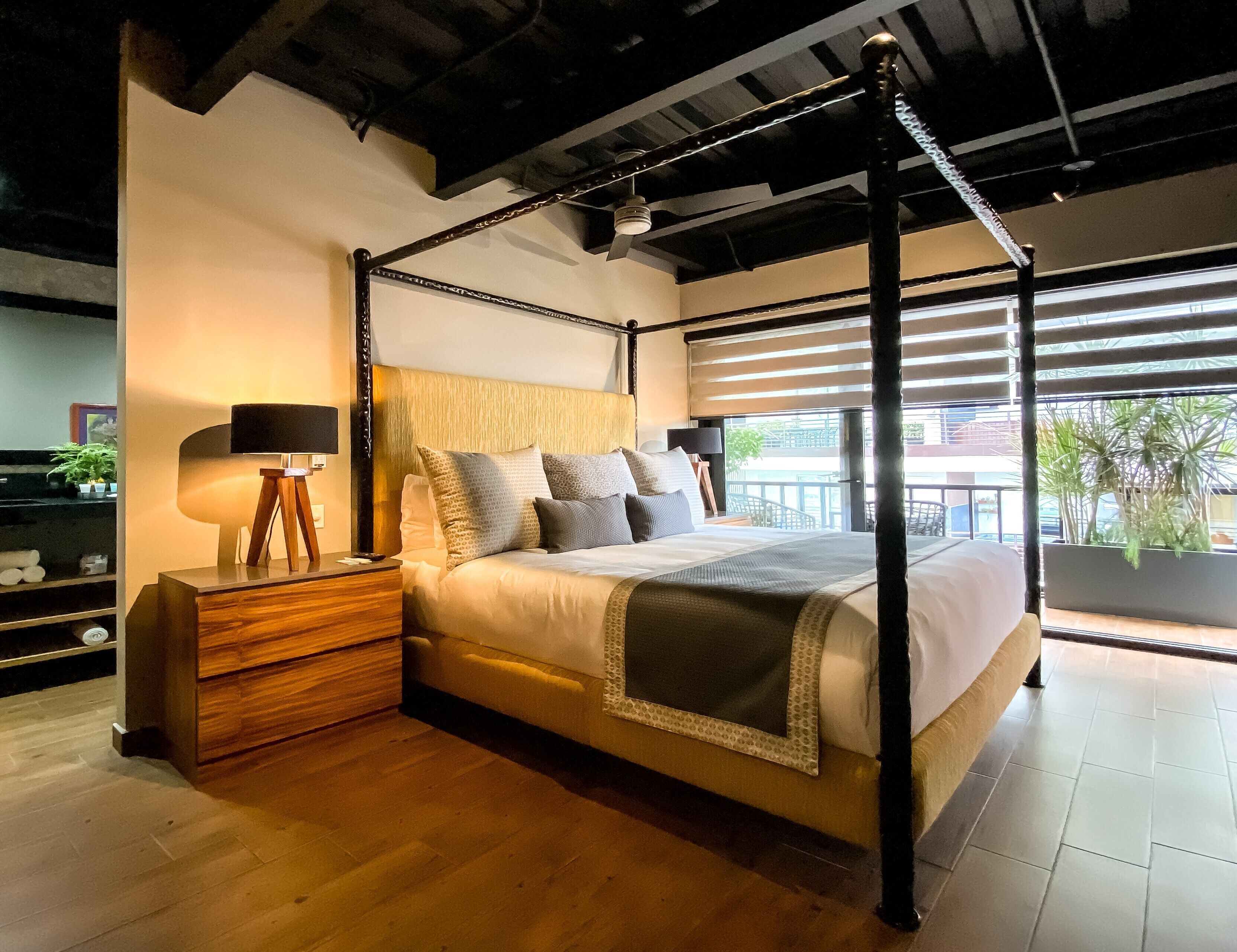 Een hemelbed met een houten hoofdeinde, een nachtkastje met een lamp en uitzicht op de omgeving door grote ramen.