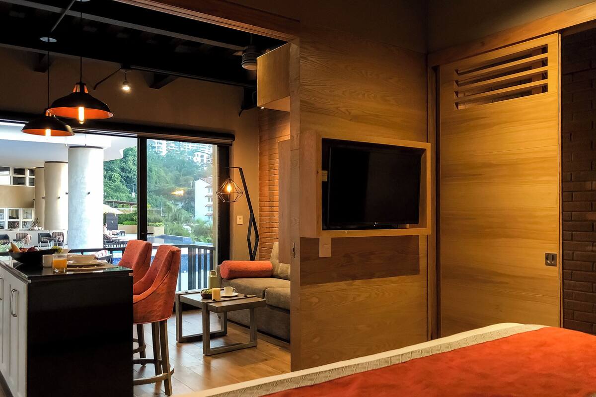 Une chambre d’hôtel moderne dotée d’un grand lit, d’un canapé, d’un coin repas avec une table et des chaises, d’un téléviseur fixé au mur et d’un balcon offrant une vue sur la ville.