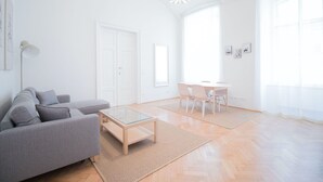 A spacious living room with a grey sofa, a glass coffee table, a dining area with a table and chairs, and large windows allowing natural light. - HeyMi Apartments Stadtpark (Vienna)