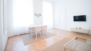 A bright, modern living room with a wooden floor, a glass coffee table, a dining area with a white table and chairs, a flat-screen TV on a white cabinet, and large windows with sheer curtains. - HeyMi Apartments Stadtpark (Vienna)