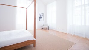 A spacious bedroom with a four-poster bed, a large window with sheer curtains, a framed picture on the wall, and a light-colored rug. - HeyMi Apartments Stadtpark (Vienna)