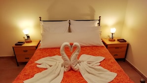 Una cama con una colcha estampada de rosas rojas, dos mesitas de noche con lámparas y una decoración de toallas con forma de cisne sobre la cama.