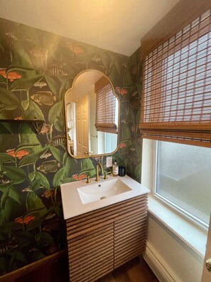 Une salle de bain avec un lavabo blanc, un miroir et une fenêtre avec des stores.