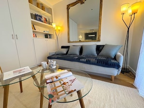 A living room with a sofa, a glass coffee table with magazines, a bookshelf, and a large mirror.