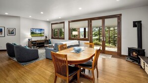 Ein modernes Wohnzimmer mit Esstisch aus Holz, bequemen Sitzgelegenheiten, einem Kamin und großen Fenstern mit Blick ins Freie.