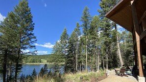 Un chalet au bord du lac, doté d’un patio couvert, d’un chemin de gravier menant au chalet et offrant une vue sur le lac, lequel est entouré de grands arbres.