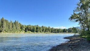 En flodbank med fri utsikt över vattnet, träden och de avlägsna bergen.