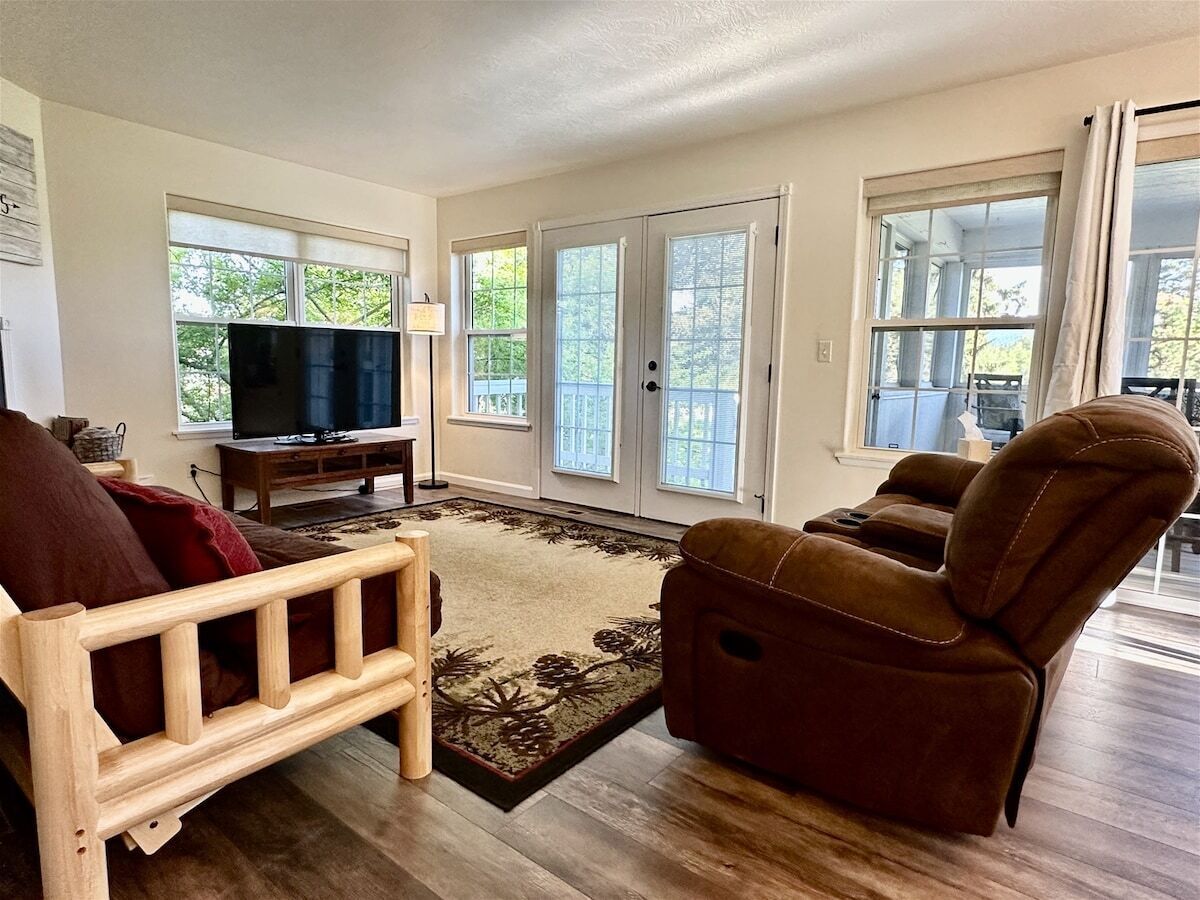 Una sala de estar con un sofá de madera, un sillón reclinable marrón, un televisor sobre un mueble bajo y un ventanal con vistas a árboles.