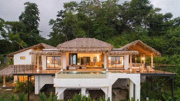Una villa moderna con piscina, construcciones de techo de paja y abundante vegetación.