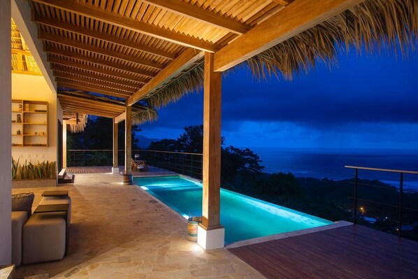 A modern outdoor pool area with a wooden pergola, stone flooring, and a view of the ocean at dusk.