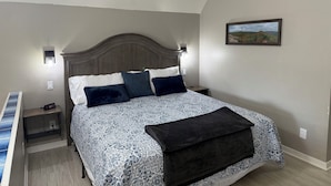 A bedroom with a large bed, a nightstand, a wall-mounted lamp, and a framed picture on the wall.