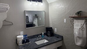 A bathroom with a granite countertop, a sink, a mirror, a towel rack, a shelf with a plant, and a coffee maker.
