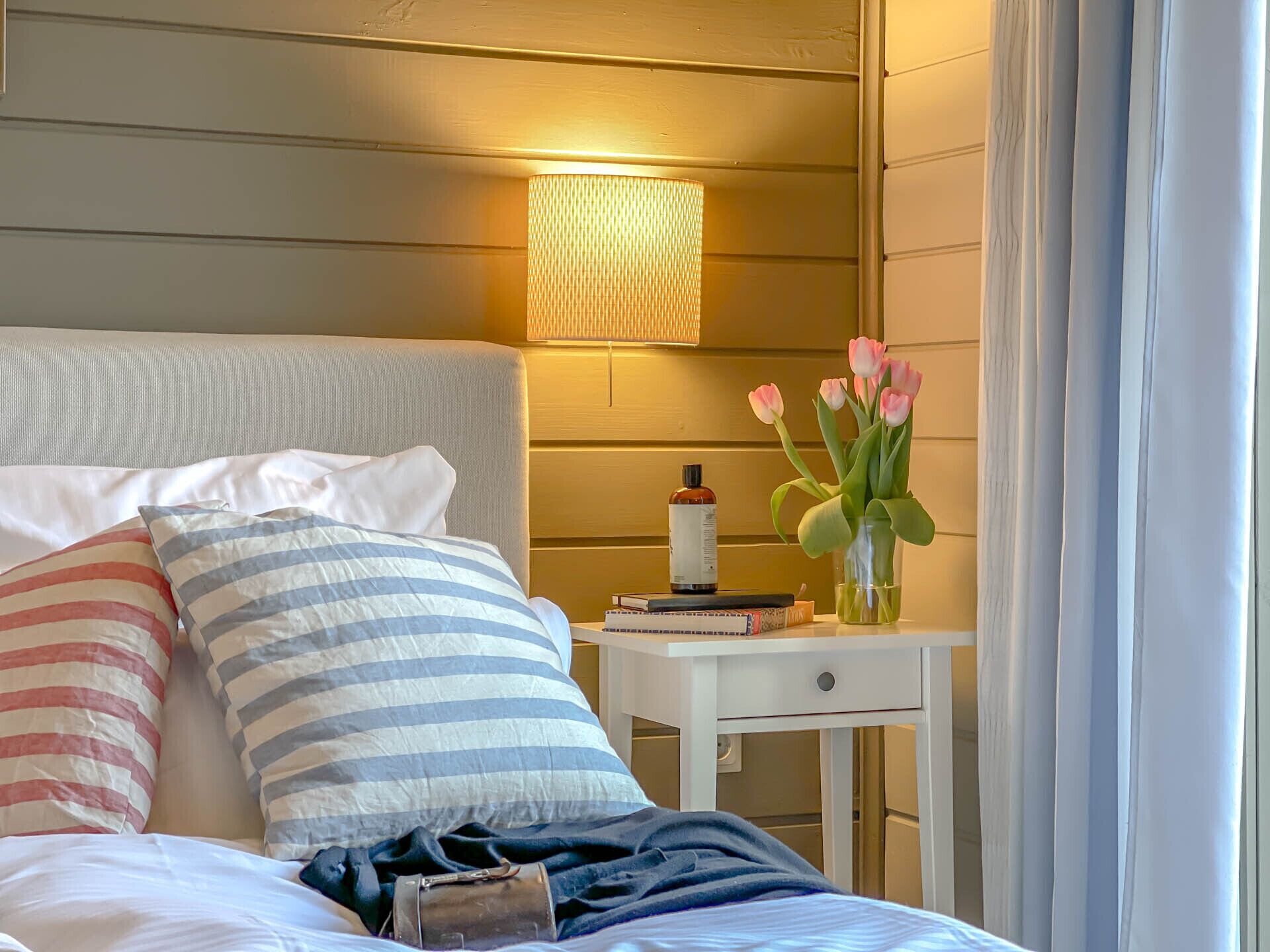 A neatly made bed with striped pillows, a bedside table with a lamp, a bottle, and a vase with flowers.