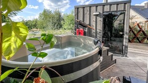 Ein Whirlpool mit Holzterrasse und Glastür, umgeben von Grünpflanzen und blauem Himmel.