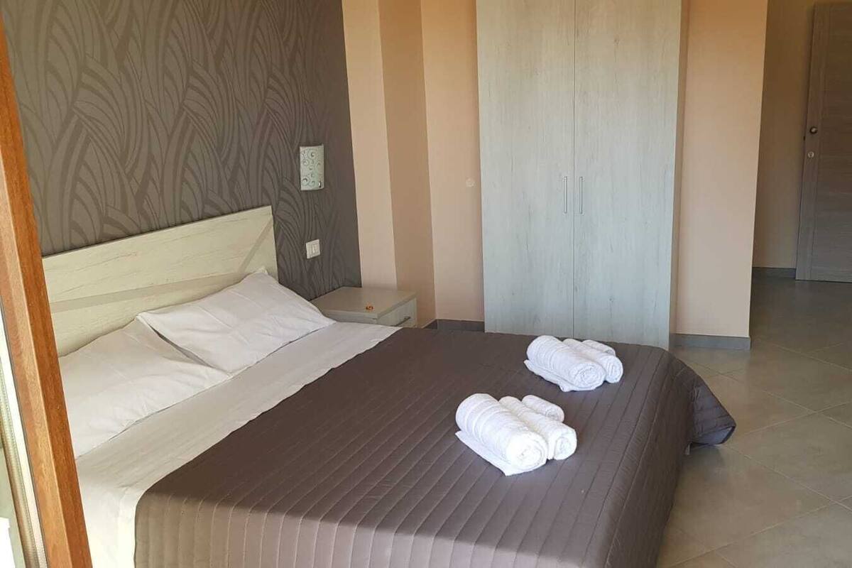 Ein ordentlich gemachtes Bett mit weißen Leinen und aufgerollten Handtüchern, ein Holz-Kopfteil, ein Nachttisch und eine schlichte Wand mit Lichtschalter.