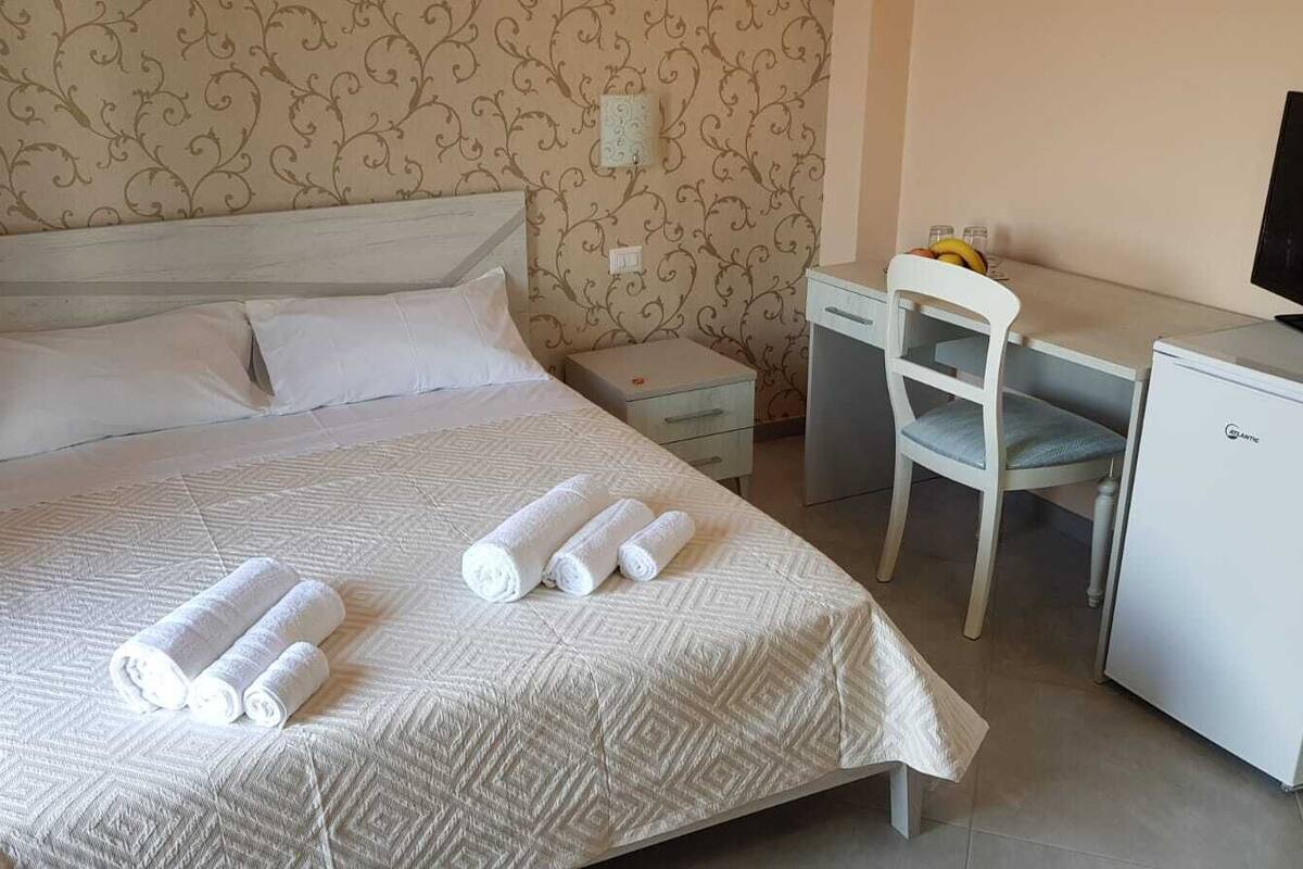 Ein ordentlich bezogenes Bett, gerollte Handtücher, ein Schreibtisch mit Stuhl und ein kleiner Kühlschrank.