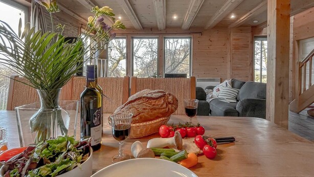 Een houten interieur met een eettafel gedekt met eten, een fles wijn en een bloemenvaas.