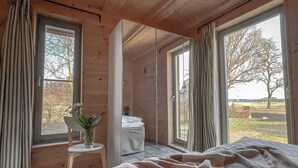 Een slaapkamer met een groot bed, een klein tafeltje met een vaas en een spiegel. Er zijn grote ramen met gordijnen en uitzicht op bomen en een hek buiten.