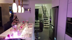 A modern kitchen with a dining area, featuring a stainless steel refrigerator, a dining table set with pink and white decor, and a staircase leading to an upper level. - Chambre Cosy (Agde)