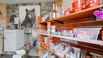 Una habitación con pared de piedra, un cuadro de un oso, un refrigerador blanco y estantes llenos de diversos objetos, incluyendo aperitivos y utensilios de cocina.