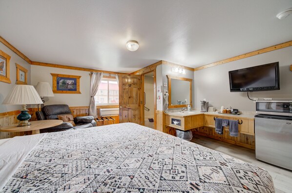 A well-lit room with a large bed, a wooden-framed desk, a chair, a television, and a kitchenette.