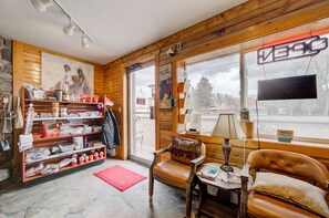 A cozy room with wooden walls, a large window, a chair, a table, and a shelf with various items.