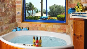 A wooden bathtub with three bottles of BREEZE beer, a window with a view of palm trees and a house, and a blue floral curtain. - The Golan Cabin (Had Nes)