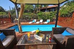 A poolside area with wicker furniture, a wooden deck, and a view of a house in the background. - The Golan Cabin (Had Nes)
