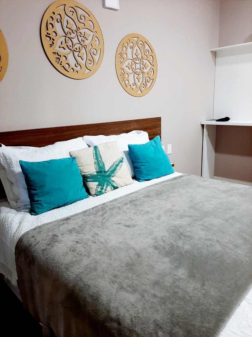 Una cama con una manta gris, almohadas blancas y dos almohadas color turquesa. Encima de la cama hay dos elementos decorativos en la pared.