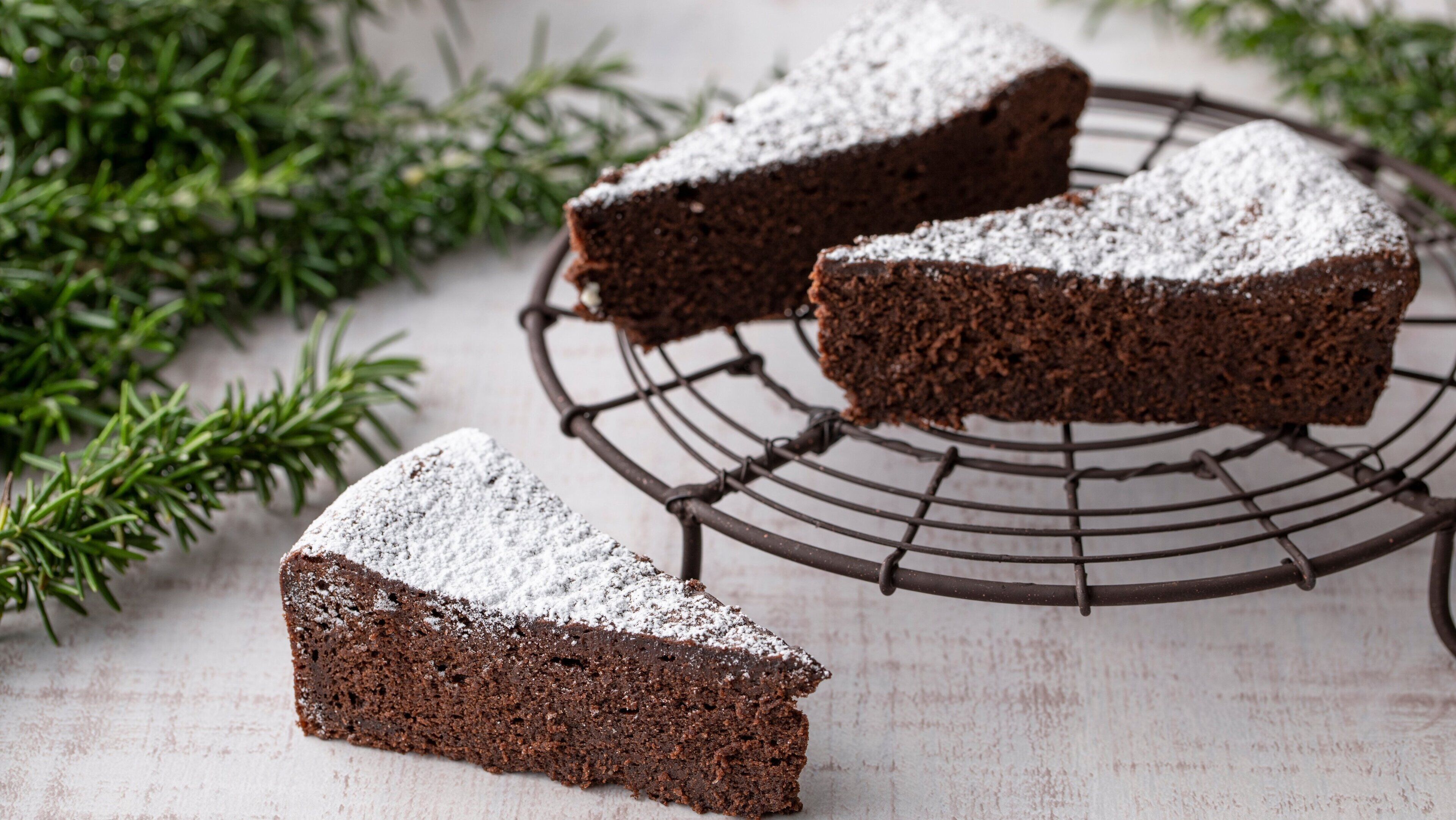A slice of cake on a wire cooling rack with powdered sugar on top, surrounded by green foliage.