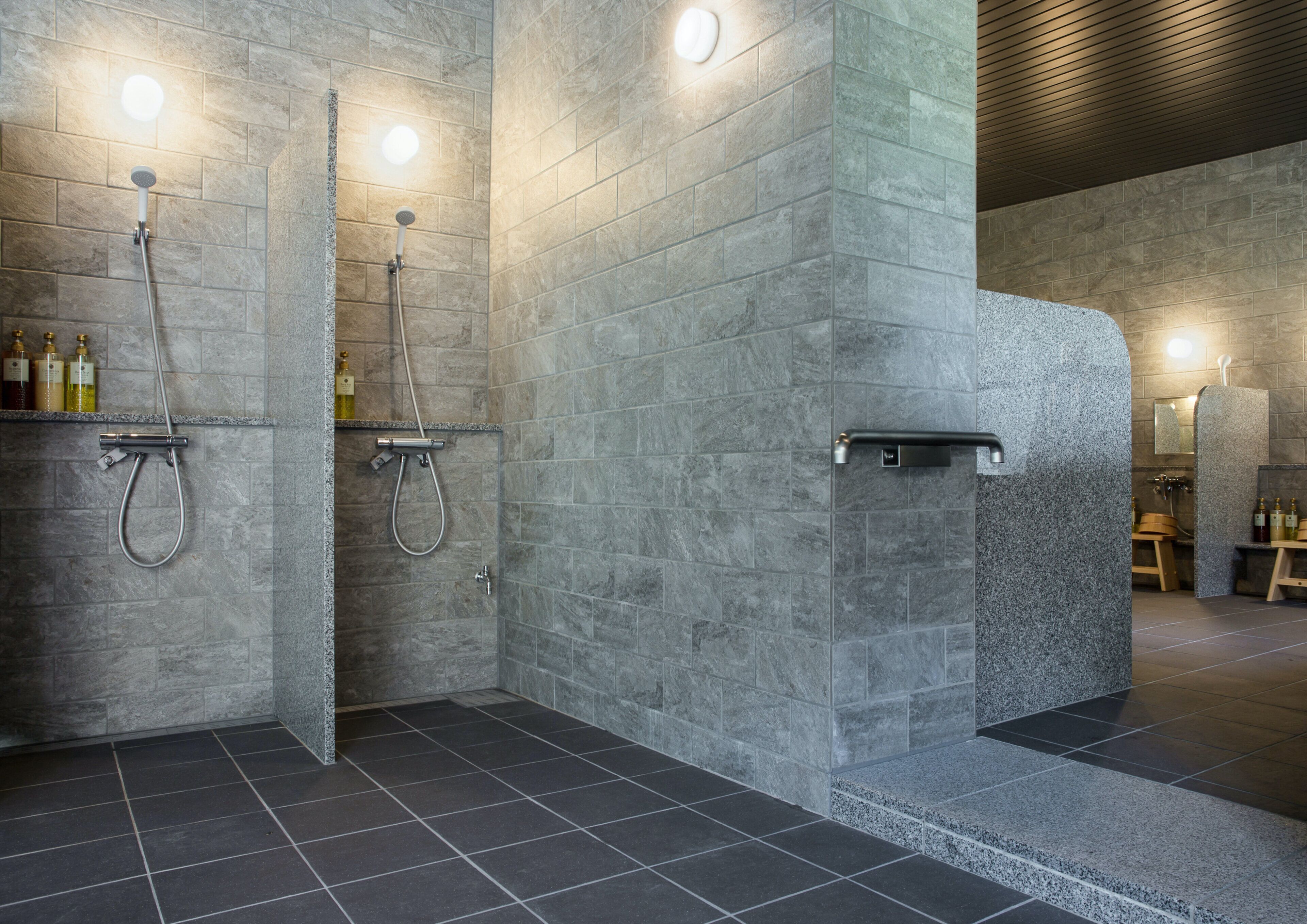 Un baño moderno con paredes de piedra gris, zona de ducha y lavamanos con artículos de aseo.