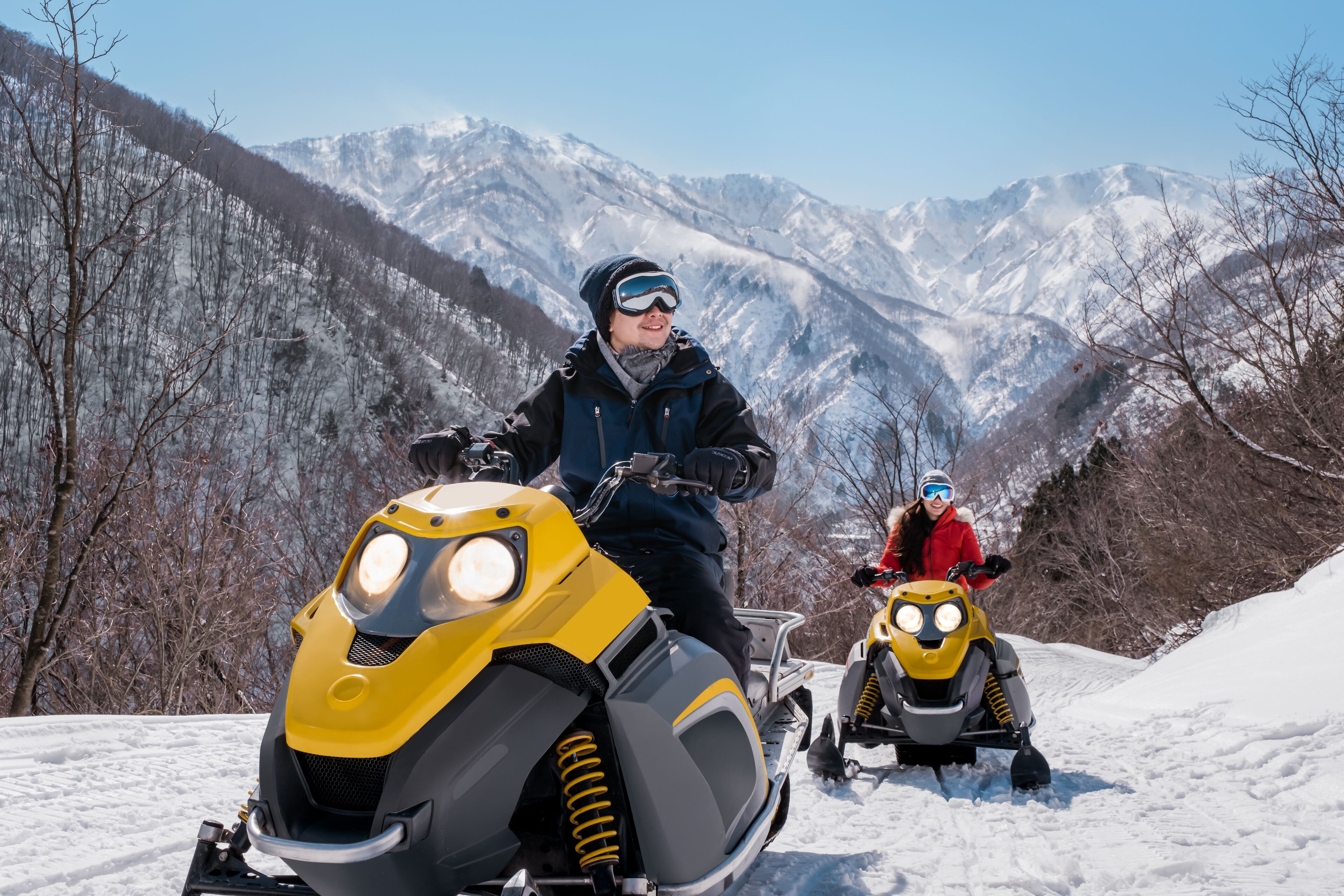 Two snowmobiles on a snowy path with mountains in the background.
