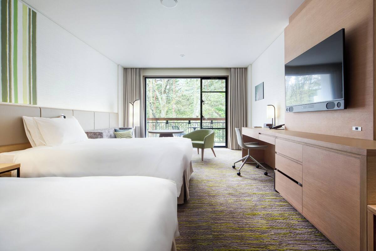 A hotel room with two beds, a desk, a chair, a TV, and a balcony with a view of trees.