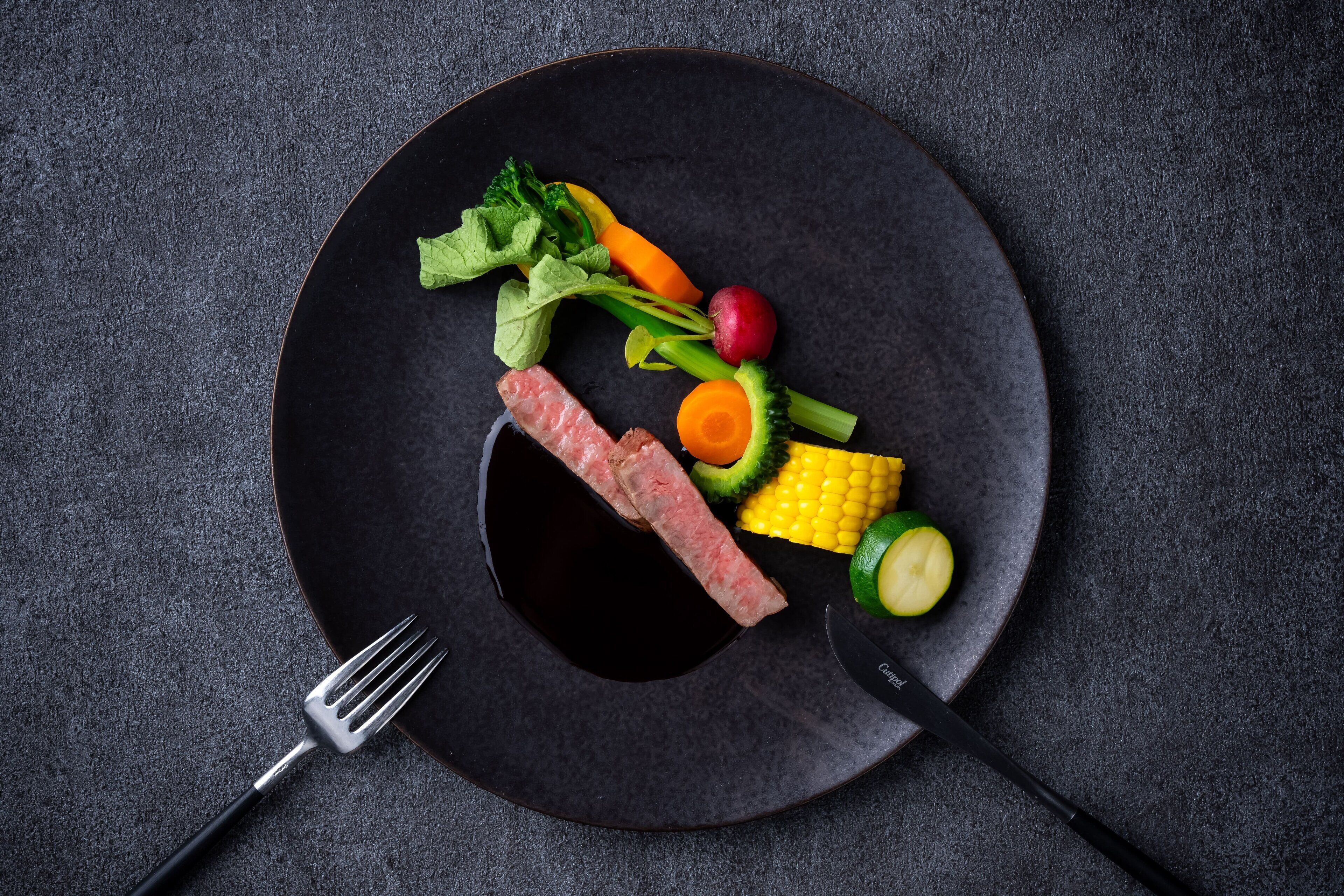 A black plate with a variety of vegetables and a piece of meat on a textured surface.