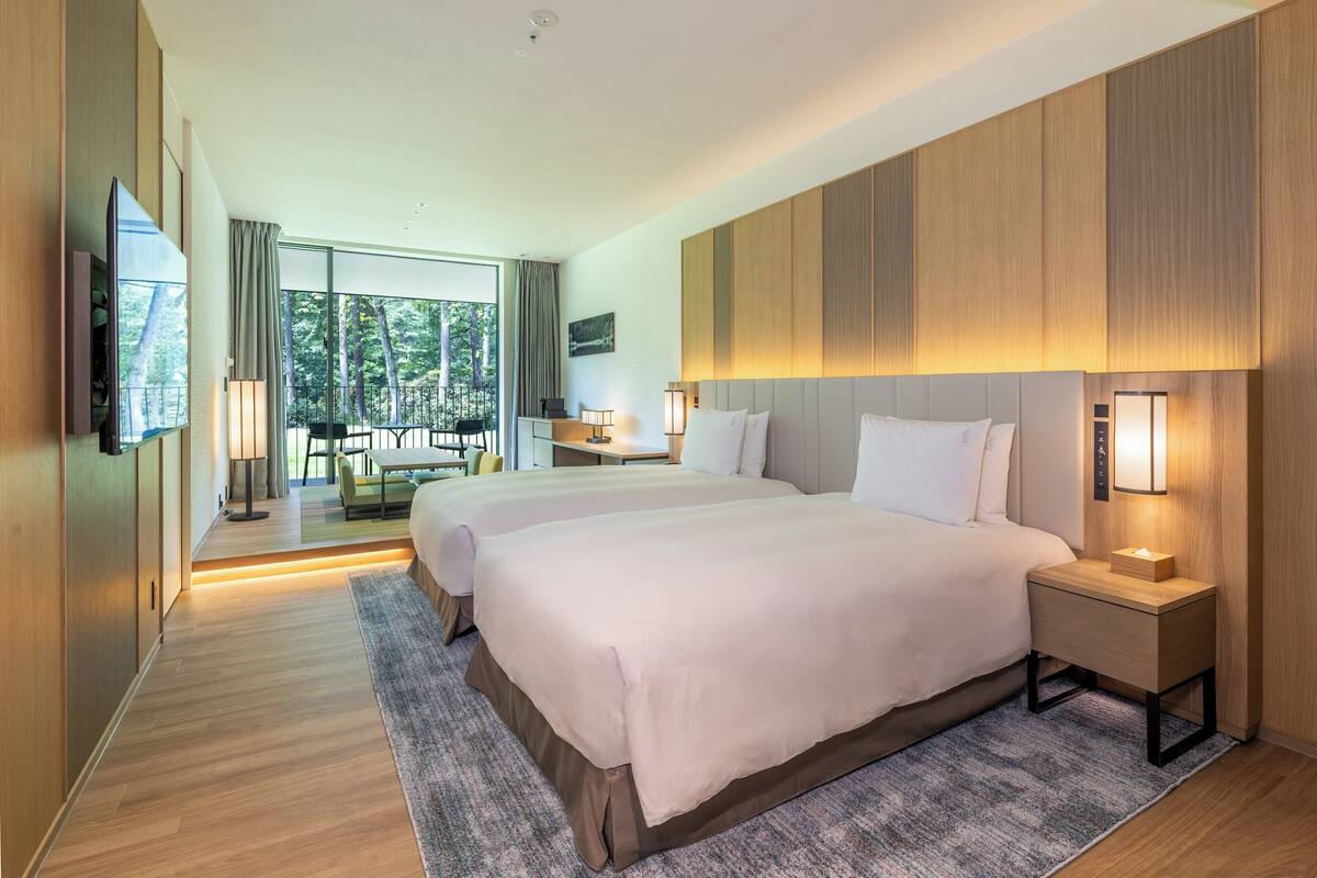 A modern hotel room with two beds, a TV, a desk, and a balcony with a view of trees.