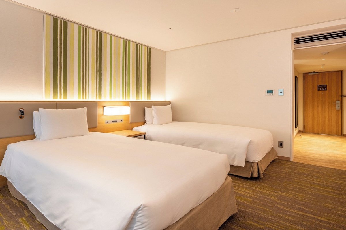 A hotel room with two beds, a bedside table, a wall-mounted light, and a door leading to another room.