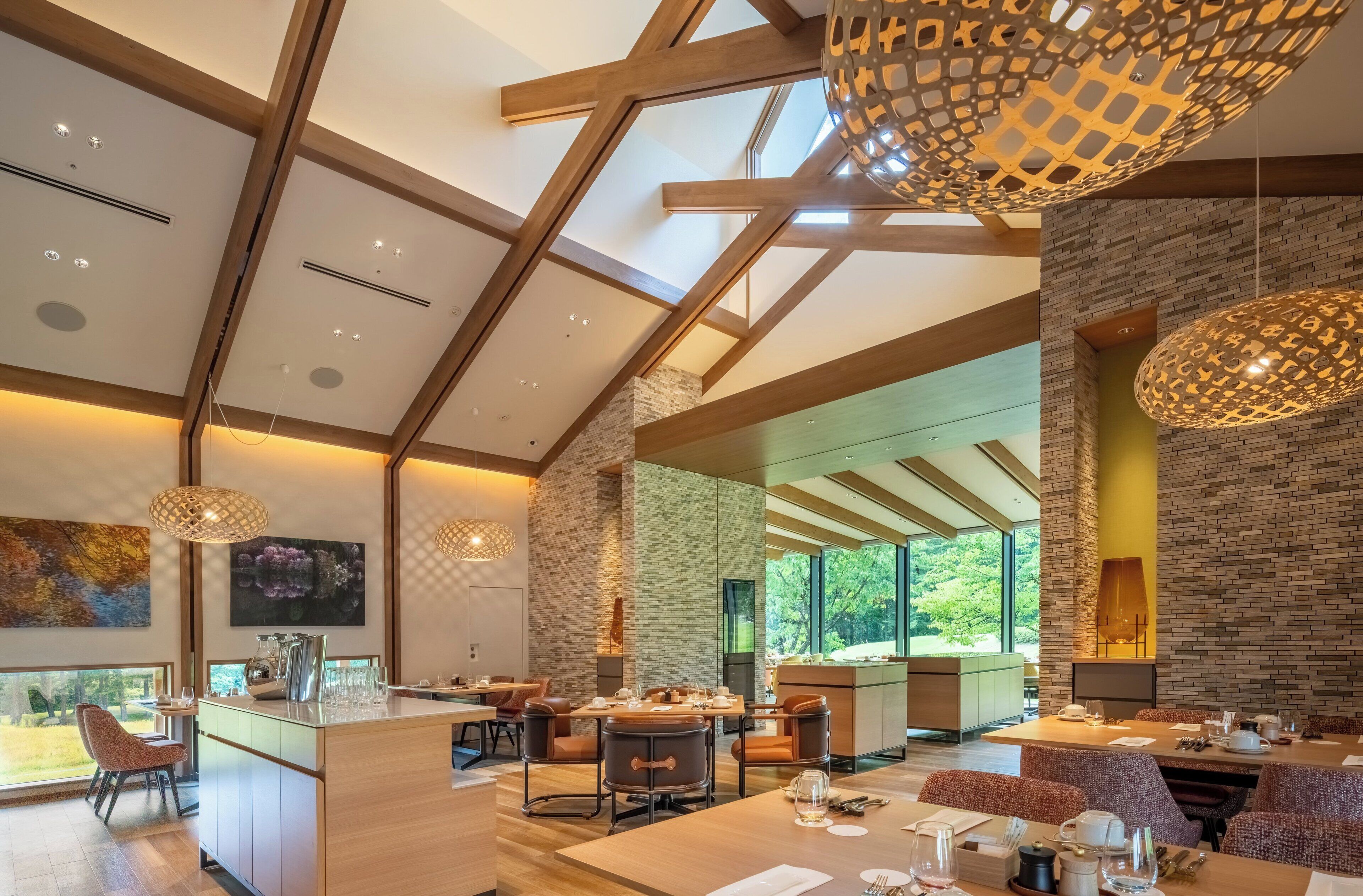 A modern dining area with a high ceiling, exposed beams, and large windows offering a view of the outdoors.