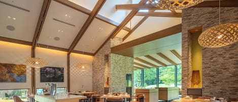 A modern dining area with a high ceiling, exposed beams, and large windows offering a view of the outdoors.