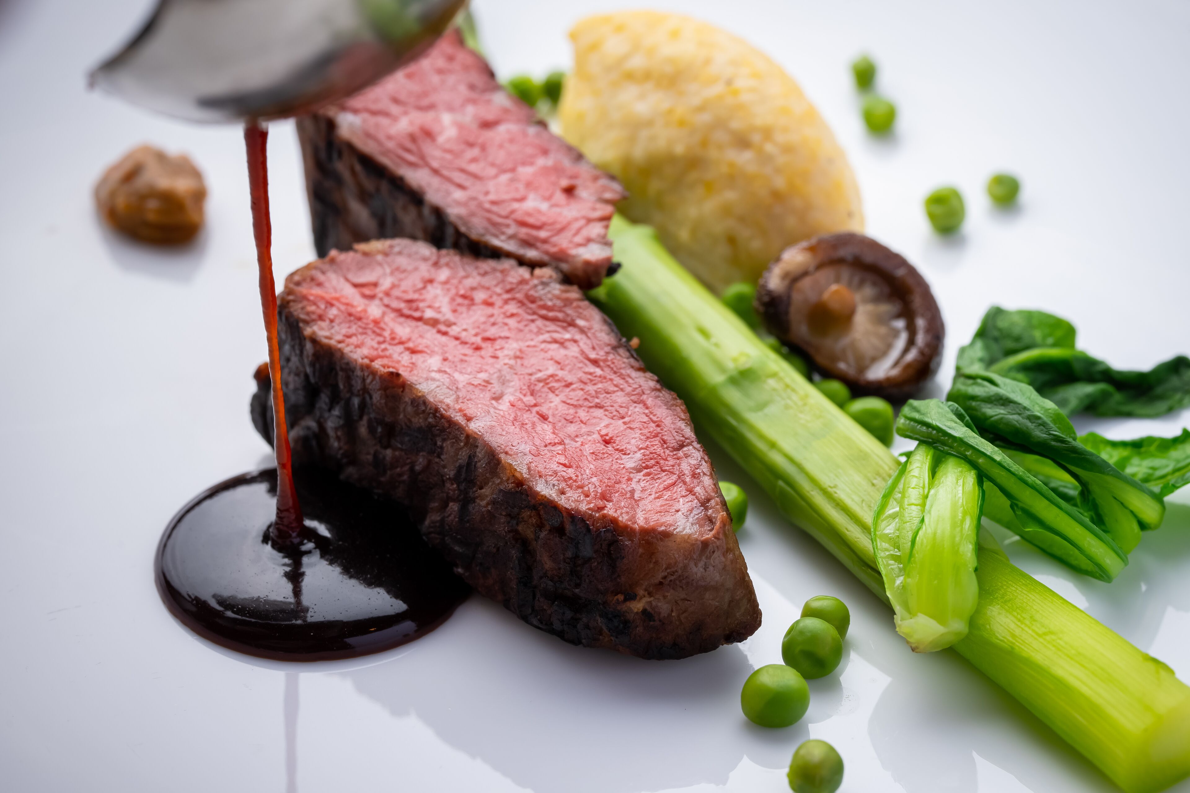 A steak with sauce, green vegetables, and a yellow item on a white plate.