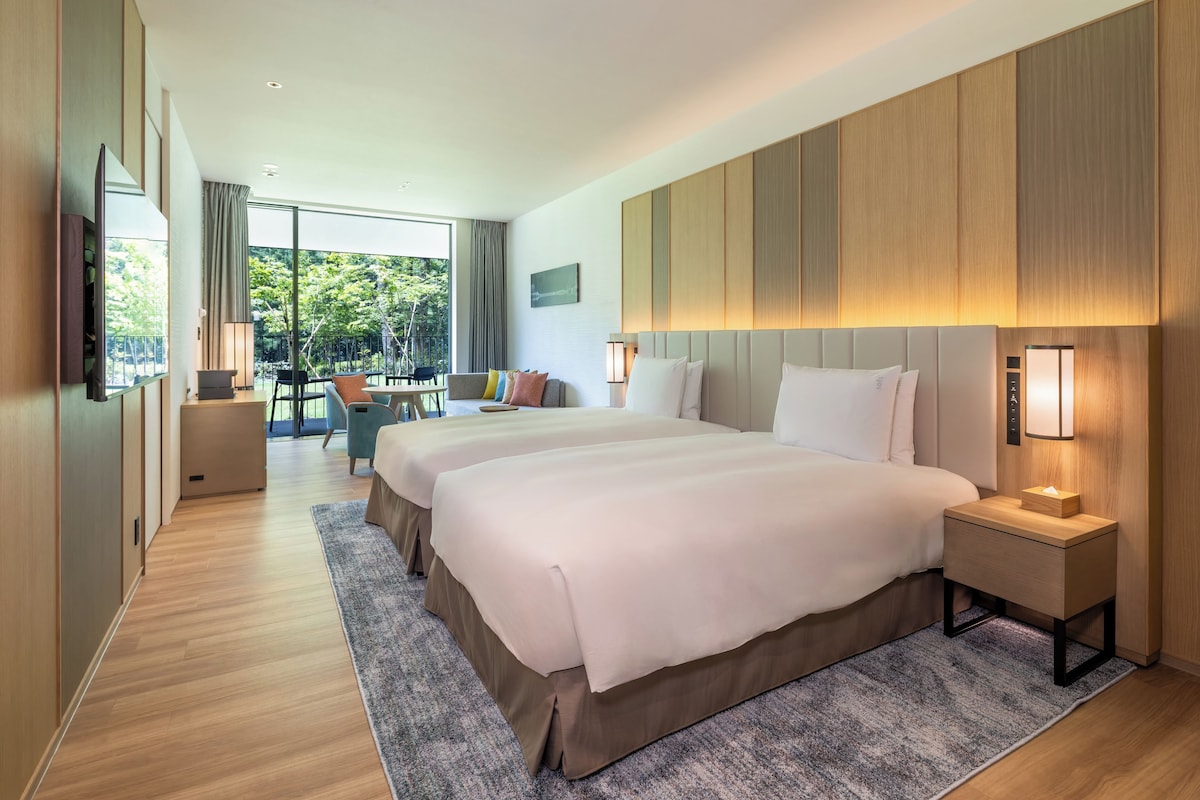 A hotel room with two beds, a sitting area with a table and chairs, a large window with a view of greenery, and a flat-screen TV mounted on the wall.