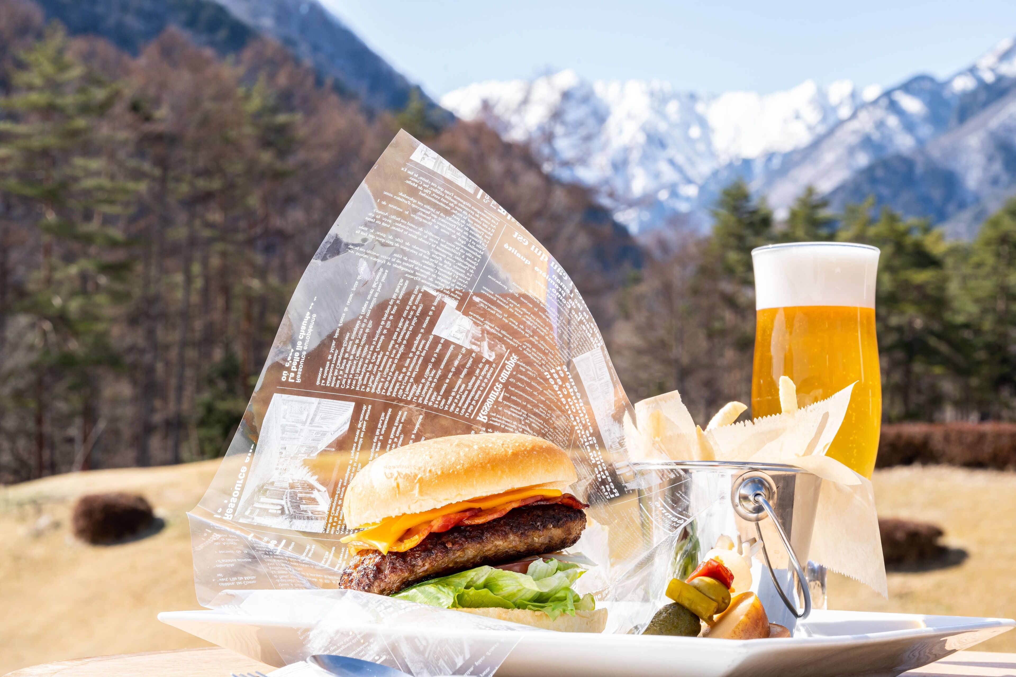 En måltid bestående av en hamburgare, pommes frites och ett glas öl, med en bergig landskap som bakgrund.