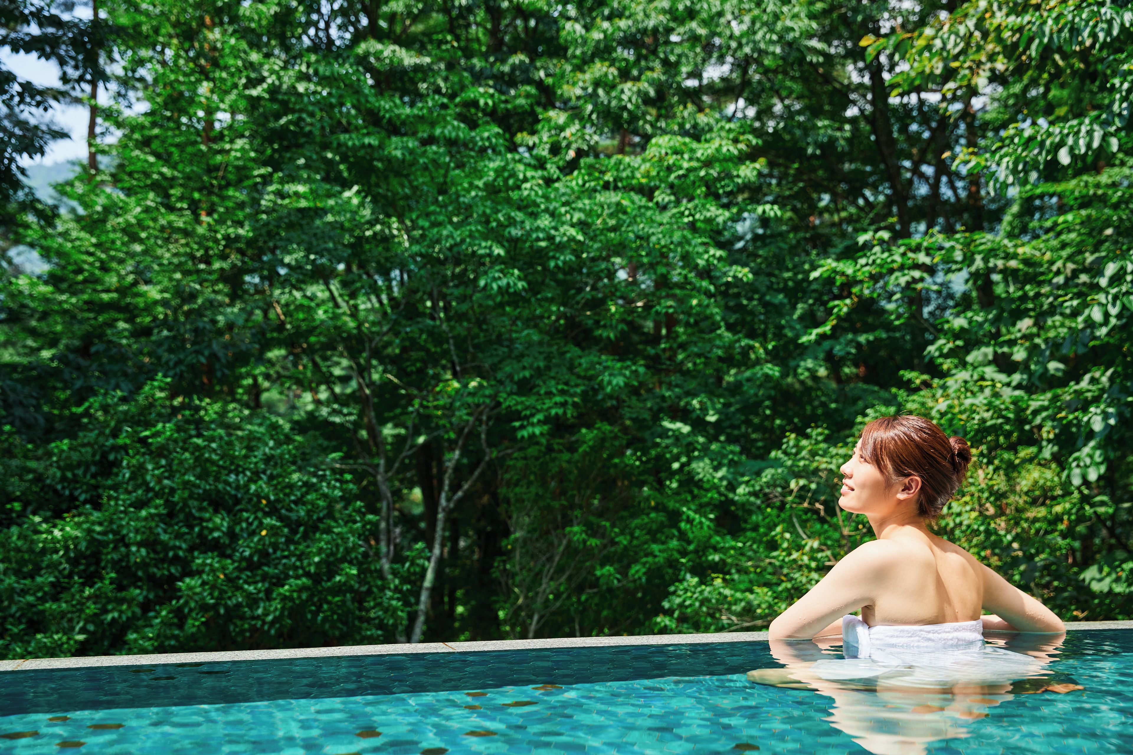 Uma pessoa em uma piscina com vista para vegetação verde exuberante ao fundo.
